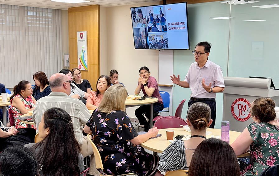 A male presenter addresses a room of seated adults during a talk on JC Academic Curriculum, with a screen displaying the presentation title in the background.